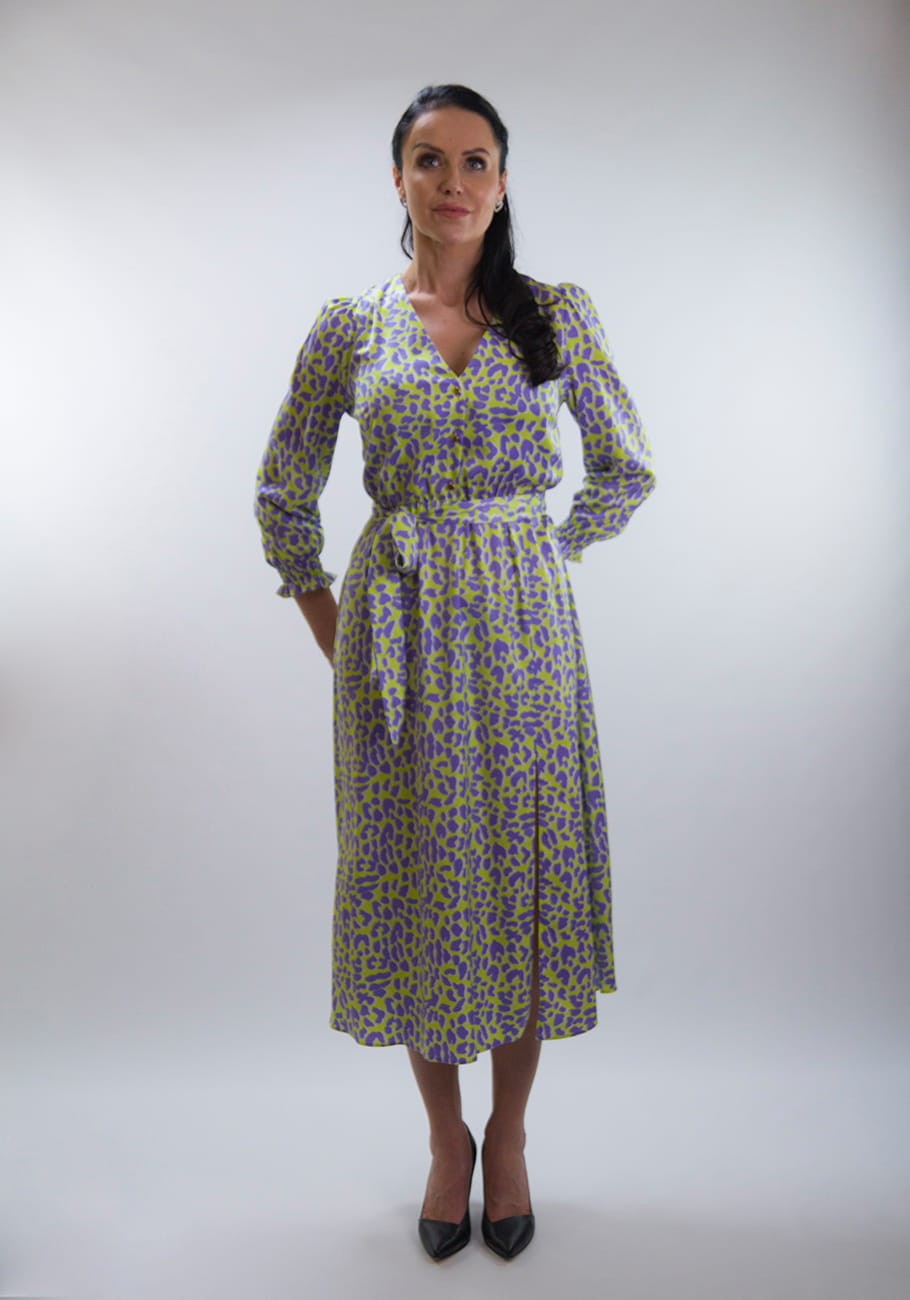 Kobieta w stonowanej pozie prezentuje się w eleganckiej sukience midi w limonkowym odcieniu z fioletowym wzorem, wykończoną długimi rękawami i dekoltem w serek. Sukienka z wiązaniem w talii podkreśla sylwetkę.