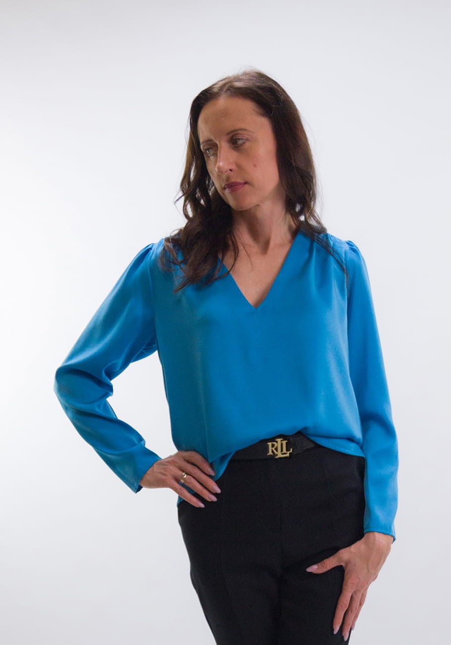 Kobieta w eleganckiej niebieskiej satynowej bluzce z dekoltem w serek, ręce luźno umieszczone na biodrach. Modelka patrzy w bok, prezentując spokojną i zamyśloną ekspresję.
