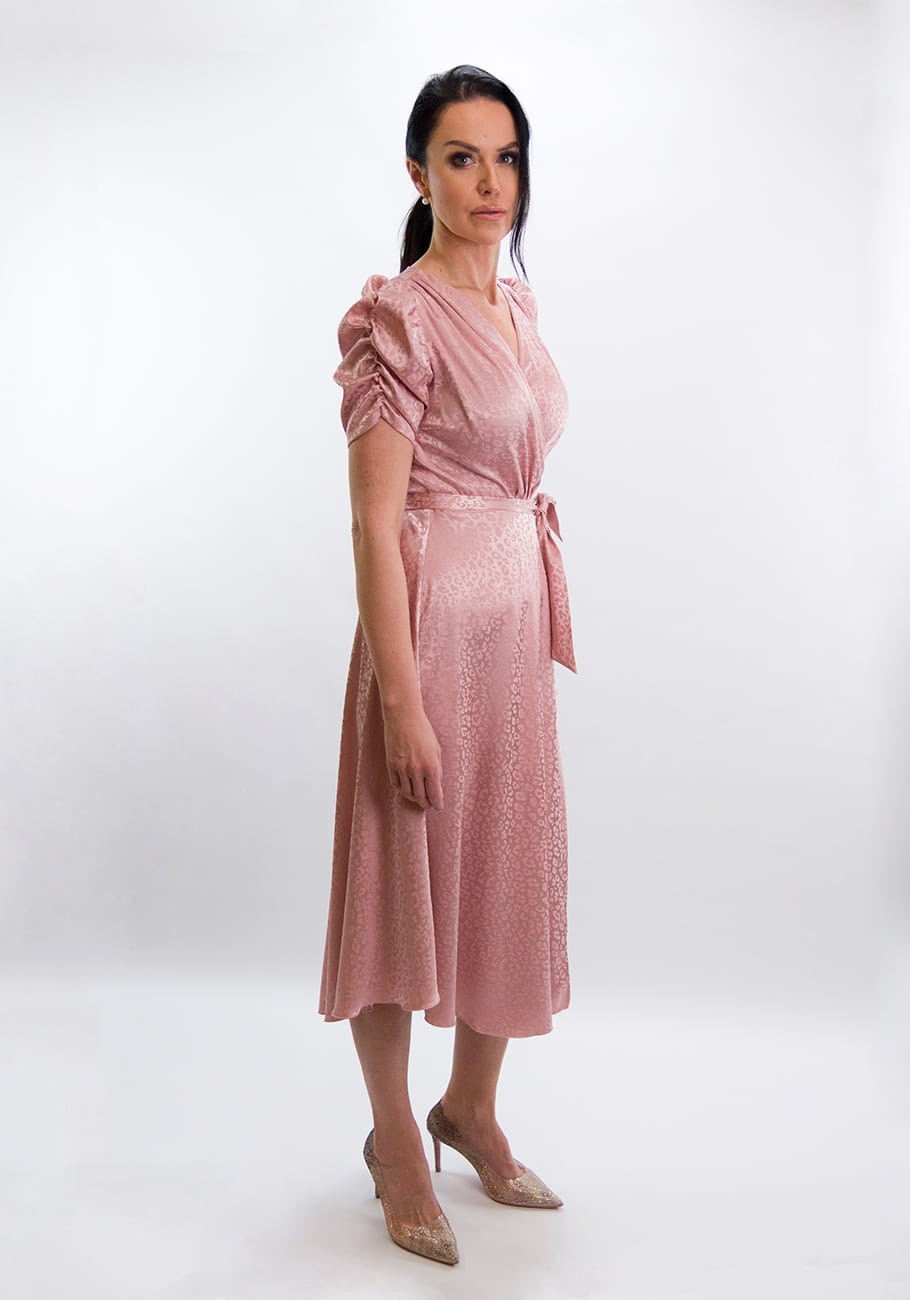 Kobieta w eleganckiej pudroworóżowej sukience midi z dekoltem kopertowym i bufiastymi rękawami, prezentująca bok sukienki, na białym tle.