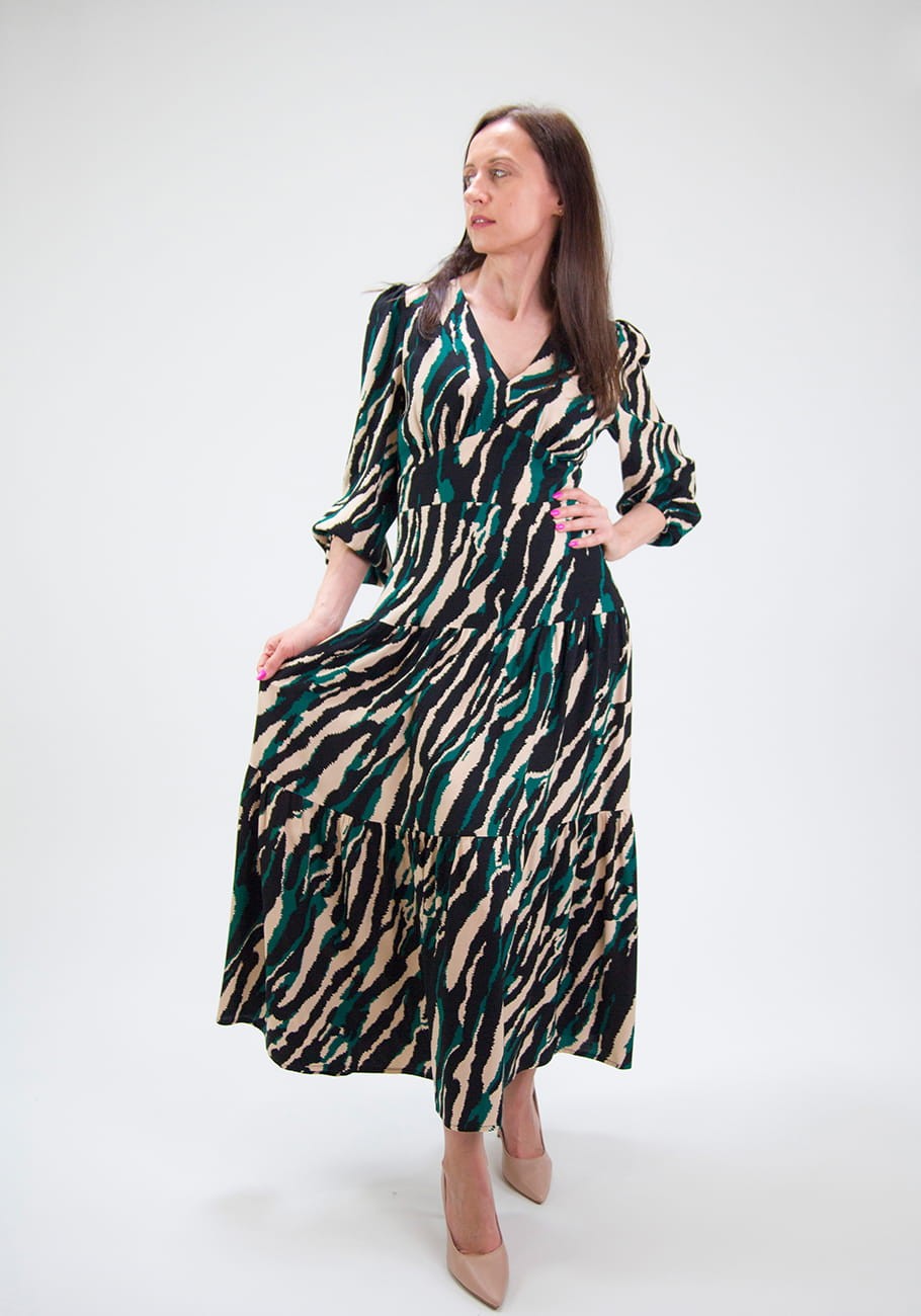 Sukienka Savannah – Midnight Jungle o długości maxi, z wiskozy, zaprojektowana z eleganckim wzorem zebry i długimi rękawami, subtelnie podkreśla sylwetkę stojącej kobiety.