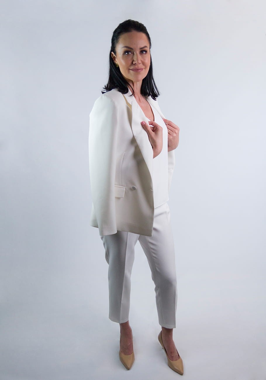 Kobieta w eleganckim garniturze Kamala koloru ecru od firmy STORM z zarzuconą na barki marynarką w ciekawej pozie. Dłonie trzyma na klapach marynarki. Garnitur przeznaczony na chrzest i komunię lub eleganckie spotkanie.