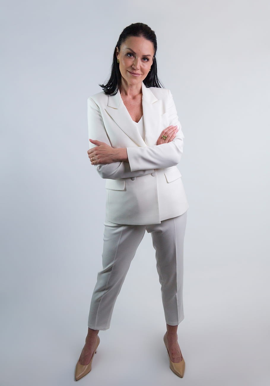 Kobieta w eleganckim garniturze Kamala koloru ecru od firmy STORM z zapiętą marynarką w ciekawej, zadziornej pozie. Dłonie trzyma skrzyżowane. Garnitur przeznaczony na chrzest i komunię lub eleganckie spotkanie.