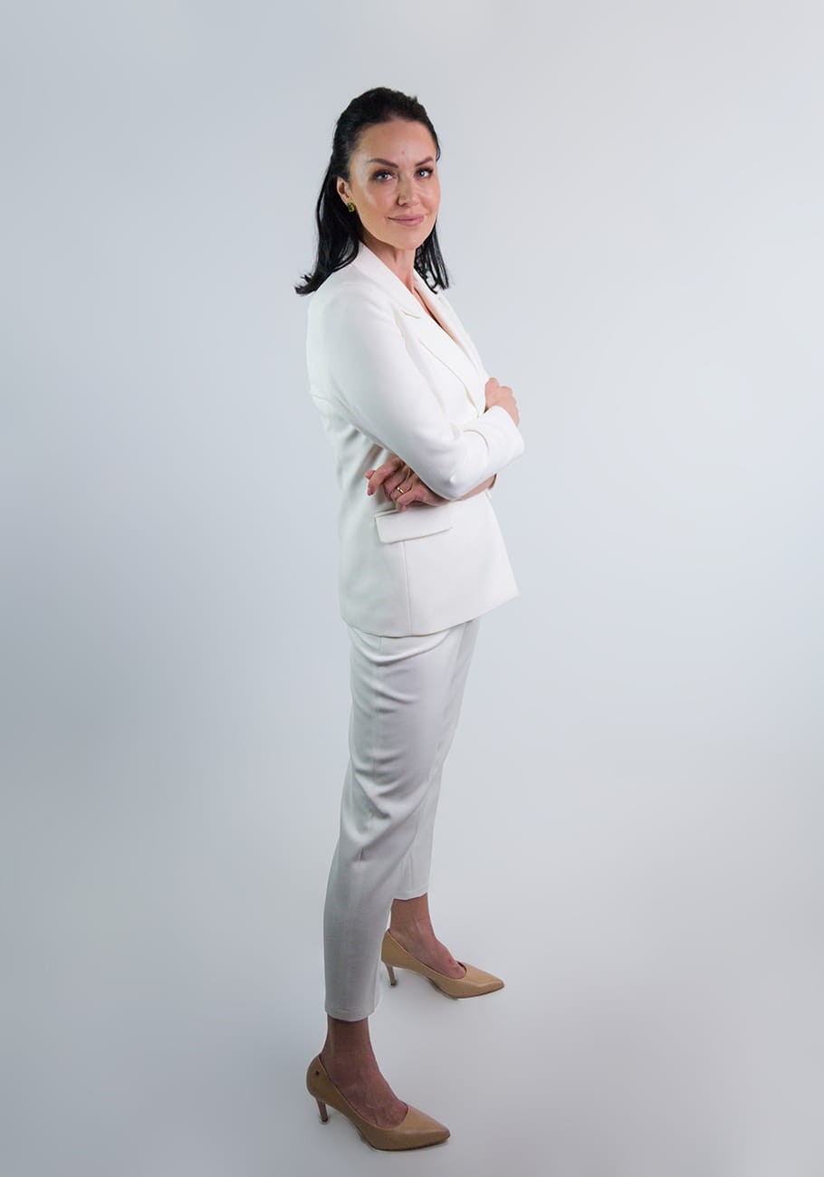 Kobieta w eleganckim garniturze Kamala koloru ecru od firmy STORM z zapiętą marynarką w ciekawej pozie. Dłonie trzyma skrzyżowane. Kobieta stoi bokiem w lekkim rozkroku. Garnitur przeznaczony na chrzest i komunię lub eleganckie spotkanie.