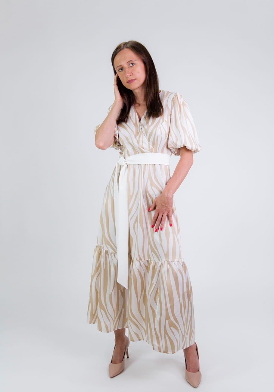 Kobieta ubrana w beżowo-białą sukienkę midi Storm Serena Beige Waves w fale z kopertową górą i bufiastymi rękawami do łokcia, trzymająca rękę przy twarzy, w beżowych szpilkach.