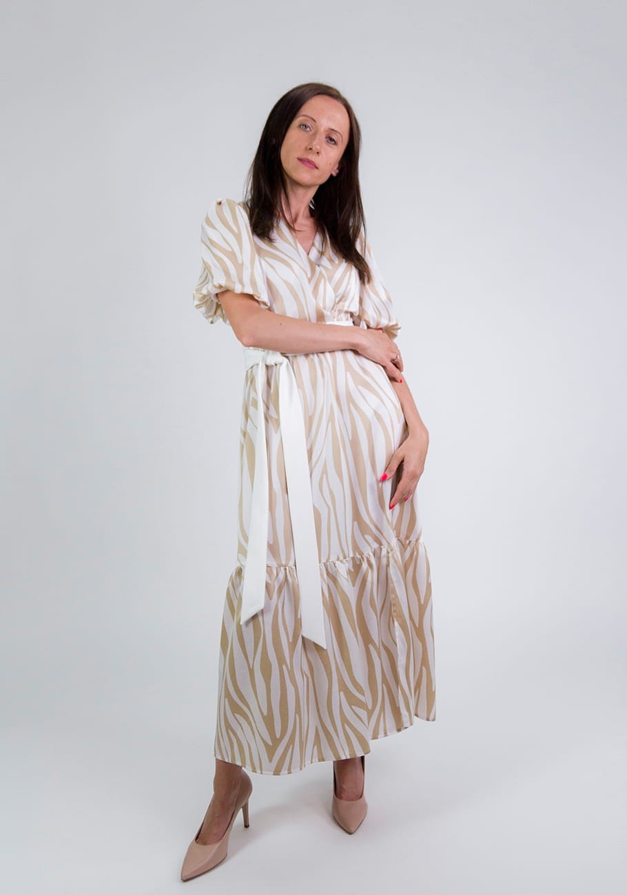 Kobieta ubrana w beżowo-białą sukienkę midi Storm Serena Beige Waves w fale z kopertową górą i bufiastymi rękawami do łokcia, skrzyżowane ręce, w beżowych szpilkach.
