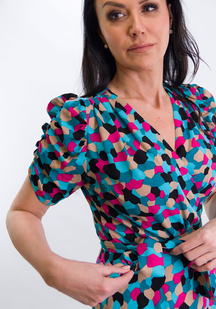 Elegancka sukienka z wiskozowego materiału, wzór w kolorowe mozaiki. Modelka stojąca, zbliżenie na górę sukienki z krótkimi rękawami bufiastymi i dekoltem w serek.