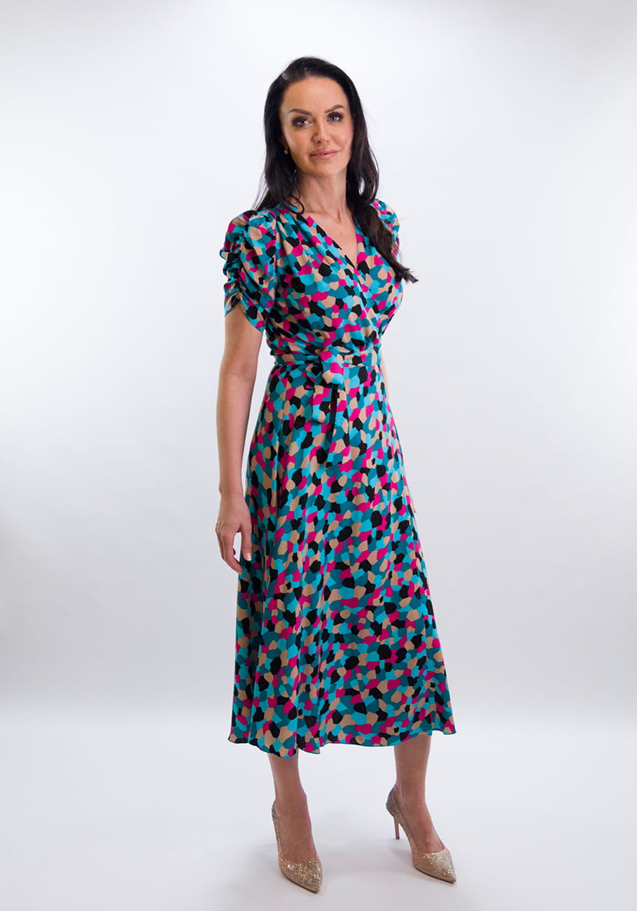 Modna sukienka midi w kolorowe, abstrakcyjne wzory, z dekoltem w serek i eleganckim paskiem podkreślającym talię. Modelka prezentuje front sukienki, dopełniony klasycznymi, złotymi szpilkami.