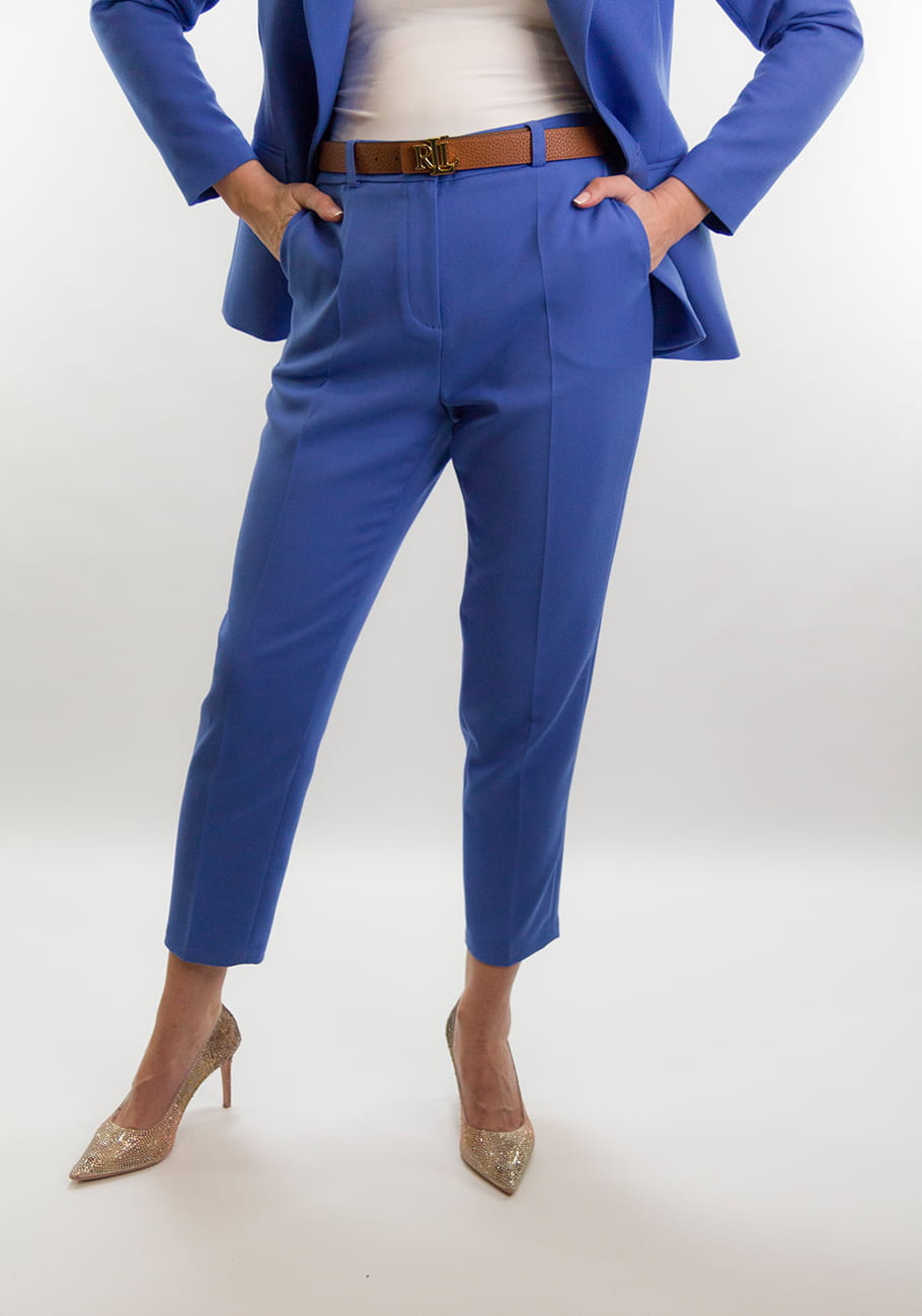 Kobieta w lekkim profilu, ubrana w niebieskie eleganckie spodnie z wysokim stanem, z brązowym paskiem z klamrą RLL, i złotymi brokatowymi szpilkami, widoczna od talii w dół, na białym tle.