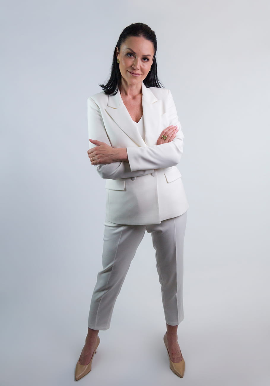 Kobieta w eleganckim garniturze Kamala koloru ecru od firmy STORM z zapiętą marynarką w ciekawej, zadziornej pozie. Dłonie trzyma skrzyżowane. Garnitur przeznaczony na chrzest i komunię lub eleganckie spotkanie.